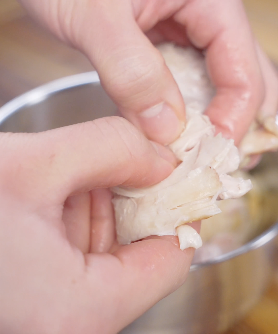 Hands shredding tender poached chicken thighs into thin strips after cooking for ginger scallion chicken.