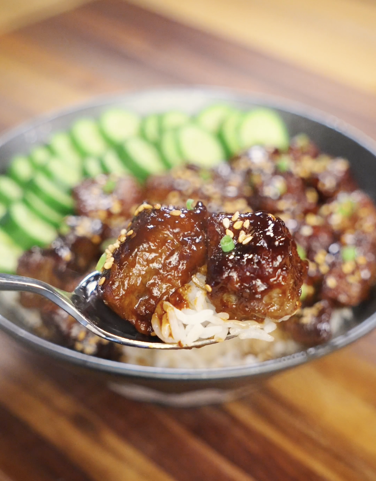 Close-up spoonful of soy garlic beef showing two tender beef cubes coated in a glossy sweet and savory soy garlic glaze, lifted over steamed white rice with sesame seeds and sliced cucumbers in the background.