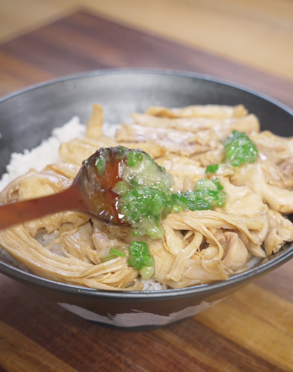 Spoonful of glossy ginger scallion oil being drizzled over shredded poached chicken on top of steamed rice, finishing ginger scallion chicken.