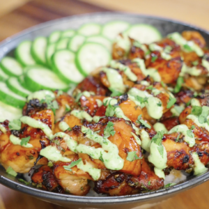 Finished Sticky Cilantro Chicken served over steamed rice with sliced cucumbers, fresh cilantro garnish, and cilantro cream drizzle in a bowl on a wooden countertop.