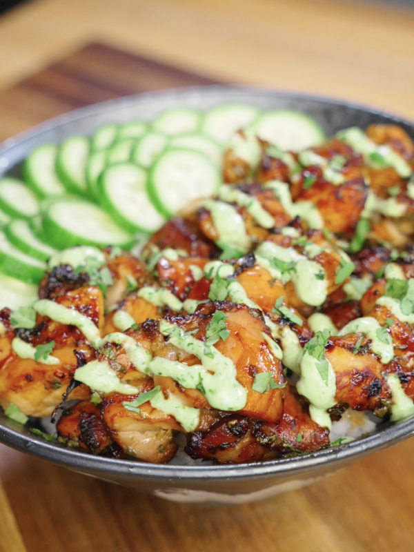 Finished Sticky Cilantro Chicken served over steamed rice with sliced cucumbers, fresh cilantro garnish, and cilantro cream drizzle in a bowl on a wooden countertop.