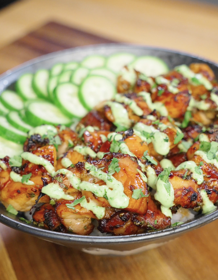 Finished Sticky Cilantro Chicken served over steamed rice with sliced cucumbers, fresh cilantro garnish, and cilantro cream drizzle in a bowl on a wooden countertop.