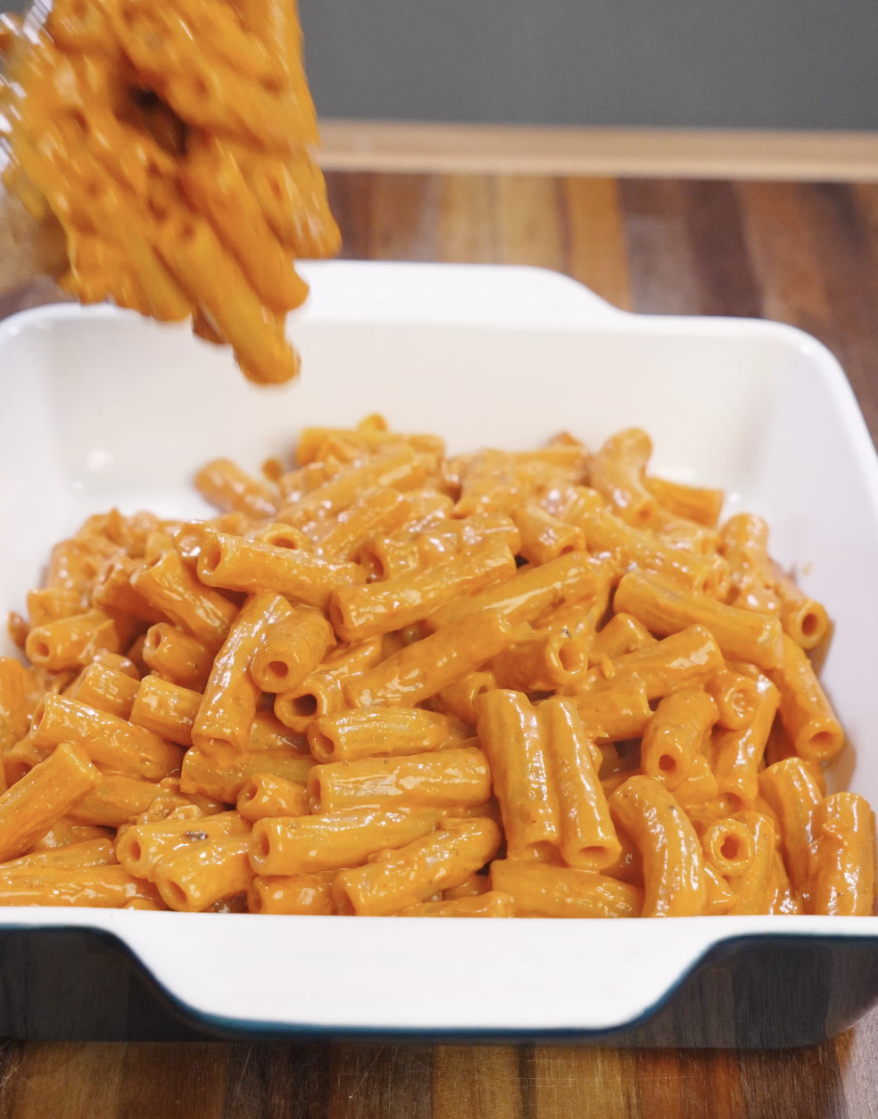 Creamy vodka pasta with rigatoni being lifted and transferred into a white oven-safe baking dish, showing noodles fully coated in a glossy tomato vodka sauce before baking.