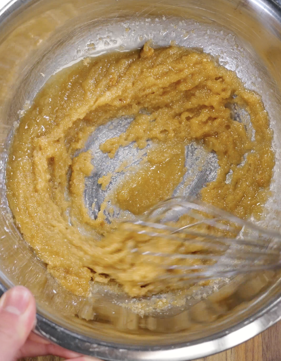 Melted butter being whisked together with brown sugar and granulated sugar in a metal bowl to create the glossy base for chewy red velvet cookies.