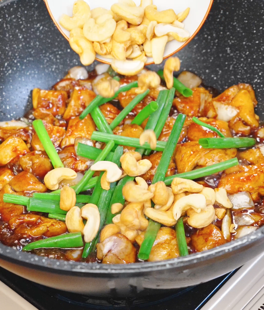 Roasted cashews and scallion greens being added to a pan of glossy honey cashew chicken during the final stir fry.