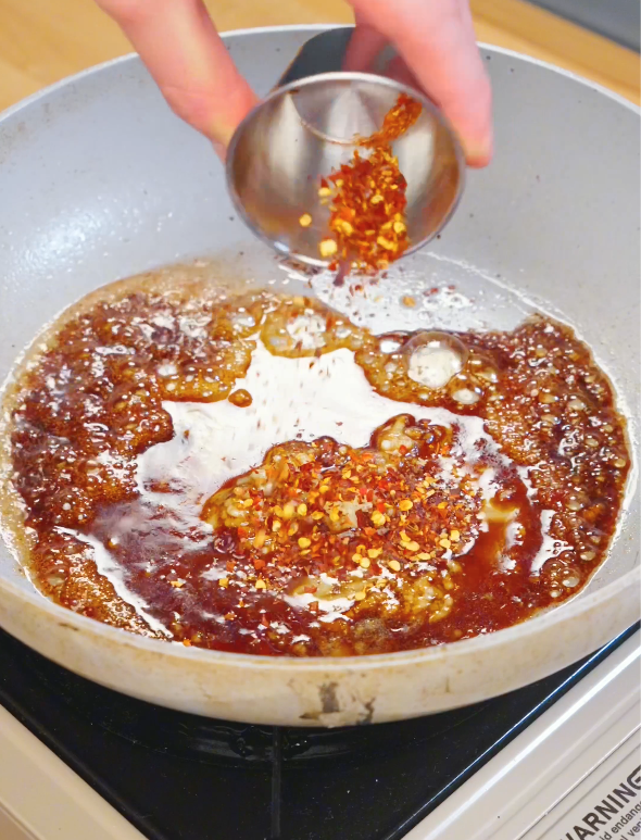 Chili flakes being poured into a pan with honey soy sauce and garlic while making the glaze for Honey Chili Chicken.
