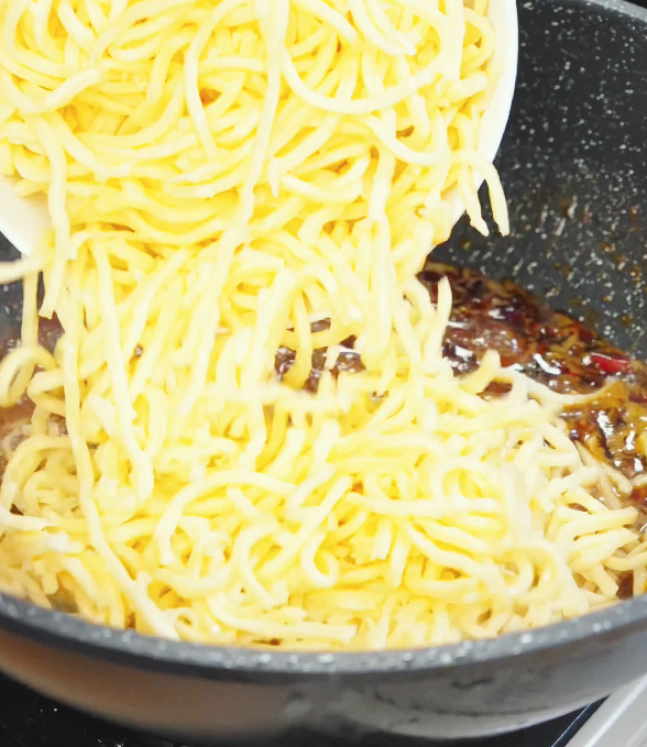 Cooked noodles being poured into a pan with chili oil garlic butter sauce to toss and coat evenly.