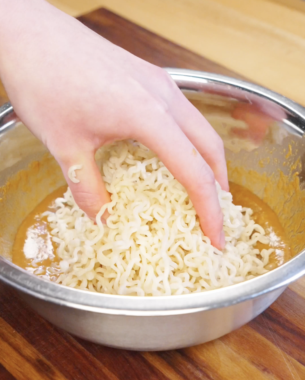 Hand placing cooked ramen noodles into a bowl of peanut butter sauce mixture.