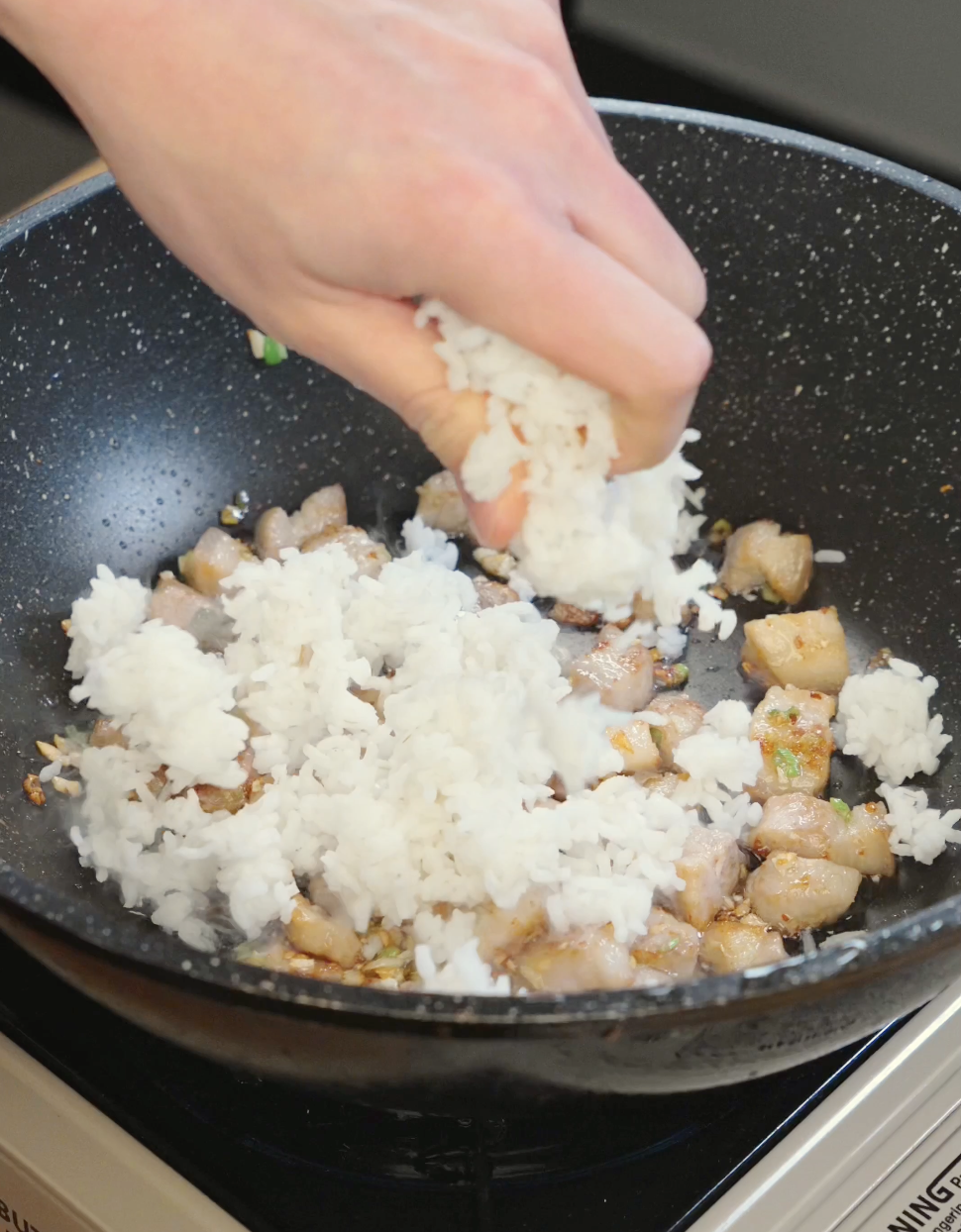 Hand breaking apart and adding cold day old rice into a pan with crispy pork belly before tossing into fried rice.