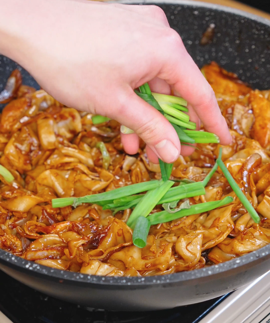 Hand sprinkling fresh scallion greens over stir fried chicken chow fun with wide rice noodles and bean sprouts.