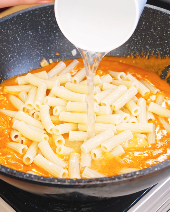 Cooked rigatoni and reserved pasta water being poured into a pan with creamy sausage sauce to finish Creamy Sausage Pasta and emulsify the sauce.