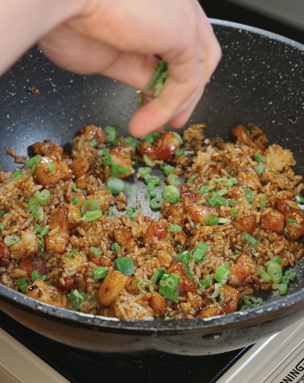 Fried rice being tossed with crispy pork belly, garlic, scallions whites, scallion greens, and sauce as the rice grains become evenly coated.