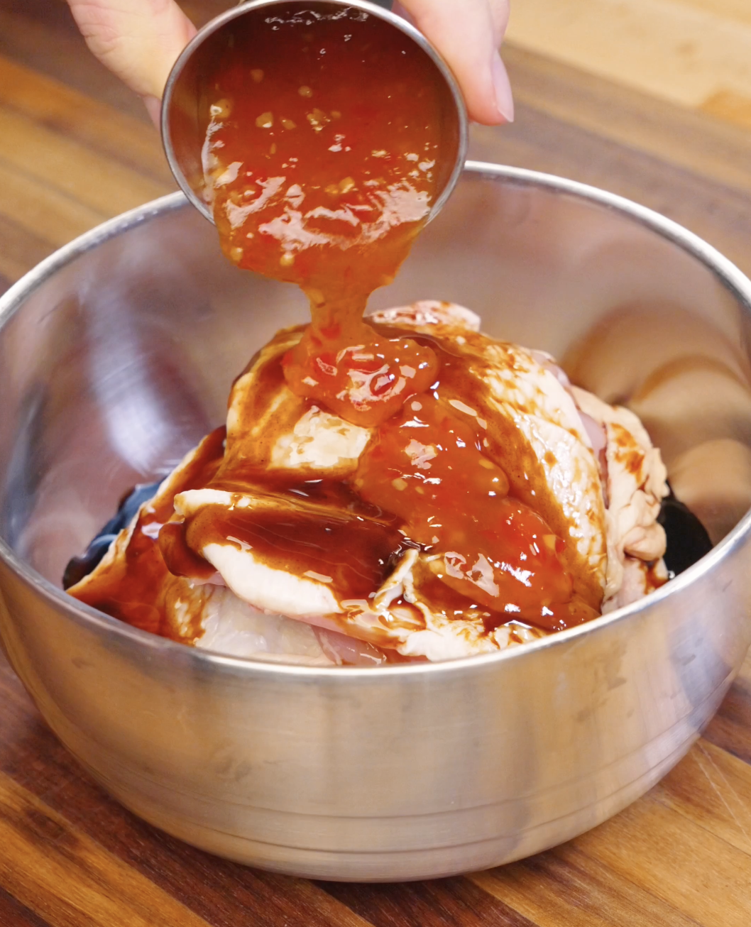 Sweet chili sauce being poured over raw chicken thighs in a bowl with soy sauce and oyster sauce marinade.