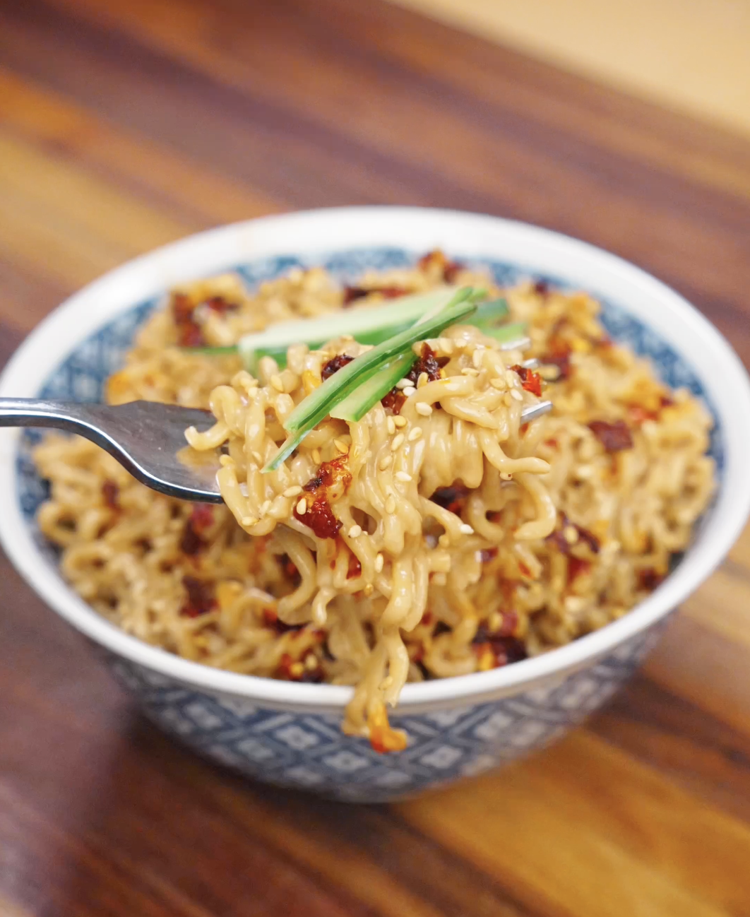 Fork lifting creamy sesame noodles topped with chili oil, sesame seeds, and cucumber garnish.
