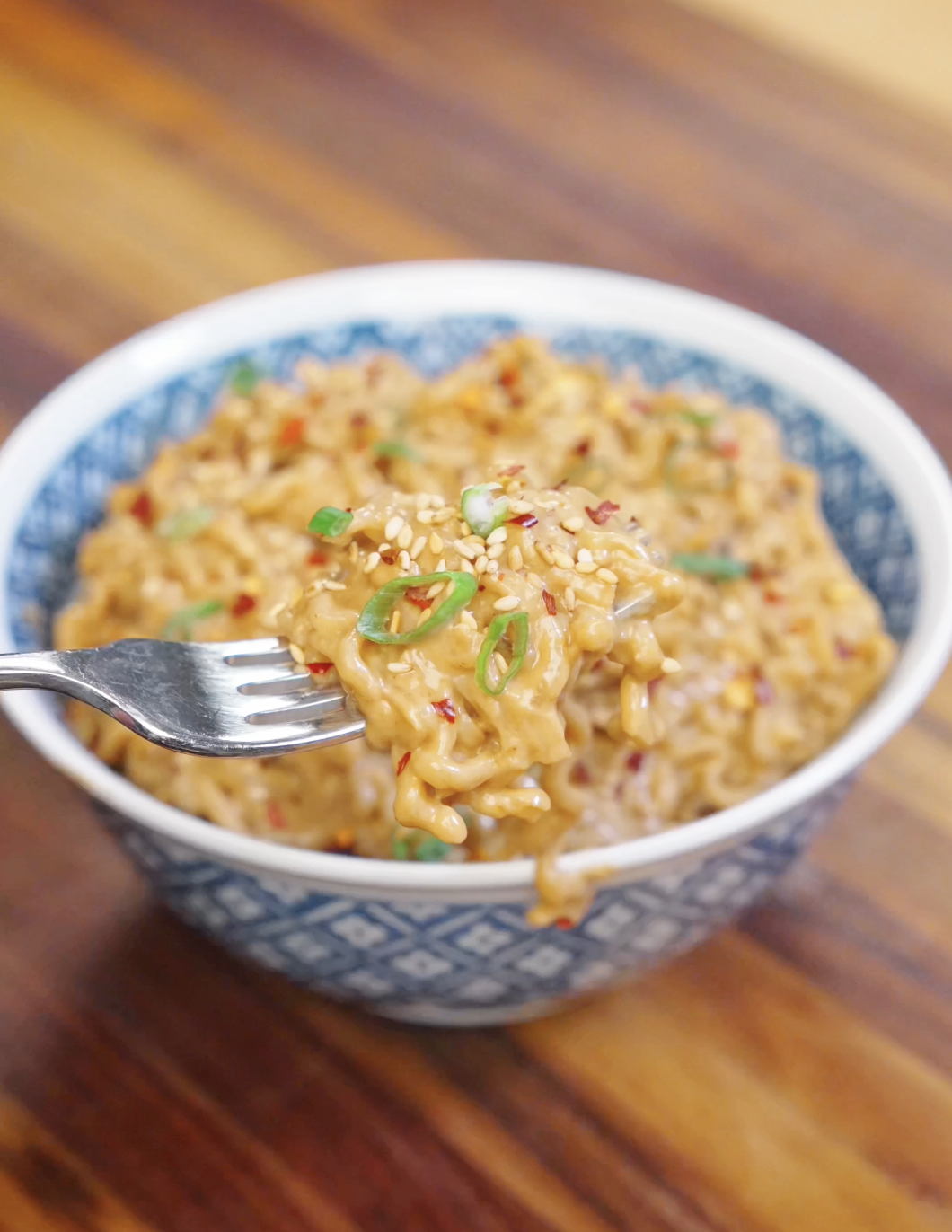 Fork lifting a bite of creamy peanut noodles topped with sesame seeds and scallions.