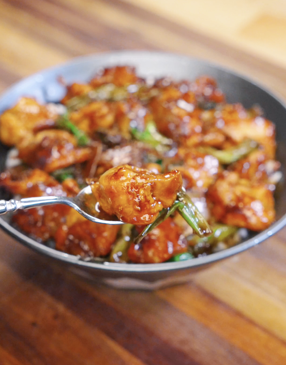 Spoon lifting a piece of glossy honey garlic chicken from a bowl of stir fry served over steamed white rice with scallions.