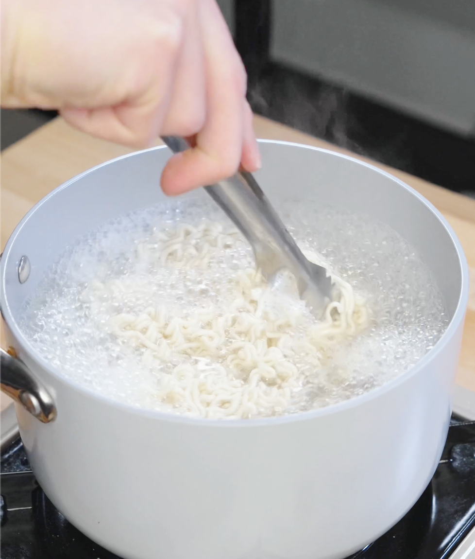Instant ramen noodles cooking in a pot of boiling water while being separated with tongs before tossing with garlic peanut butter sauce.
