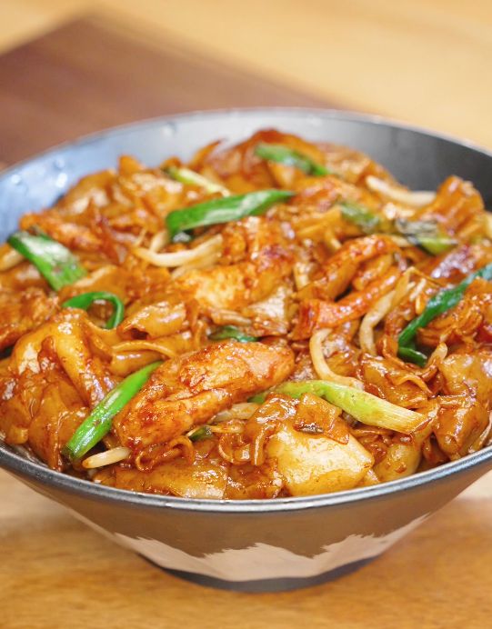 Bowl of chicken chow fun with glossy wide rice noodles, tender chicken slices, bean sprouts, onions, and scallions coated in savory soy sauce.