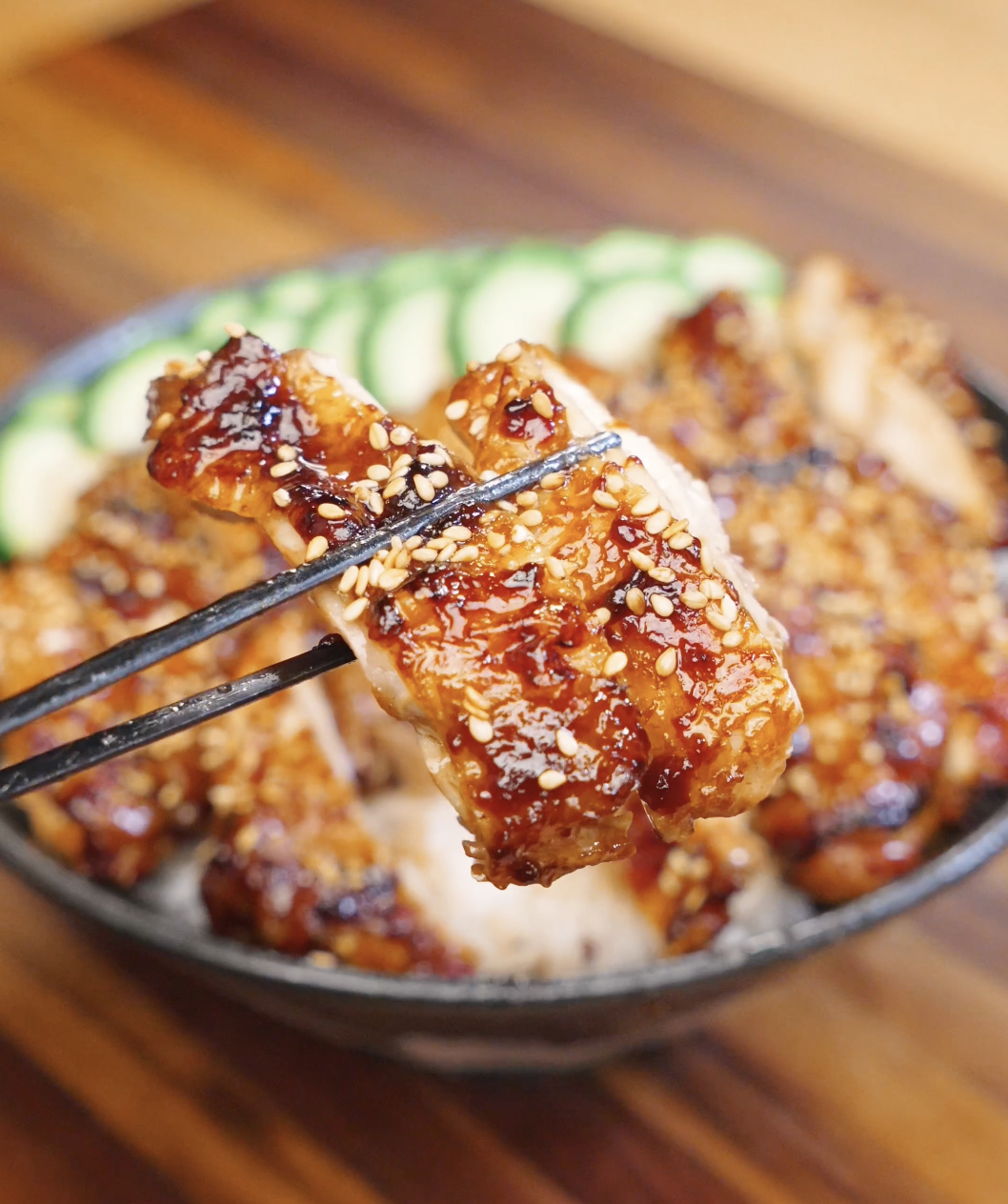 Chopsticks holding a glazed piece of Sweet Chili Chicken topped with sesame seeds over a rice bowl with cucumbers.