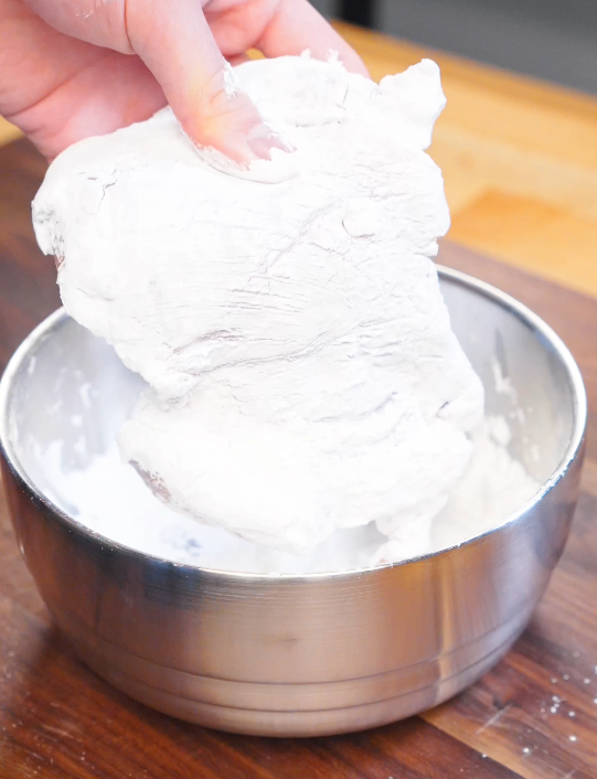 Raw chicken thigh being pressed into a bowl of cornstarch to create a crispy coating before pan frying.