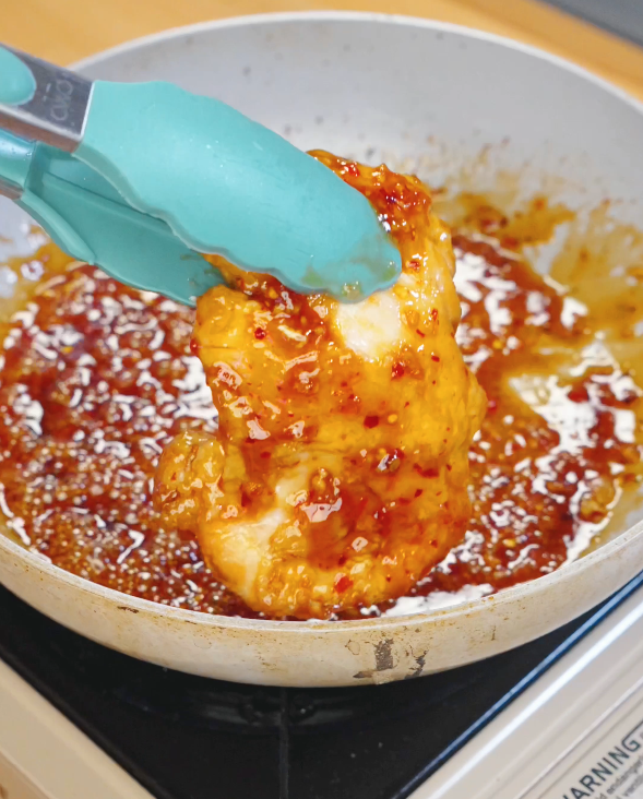 Tongs lifting crispy chicken thigh coated in thick honey chili sauce glaze inside a skillet.