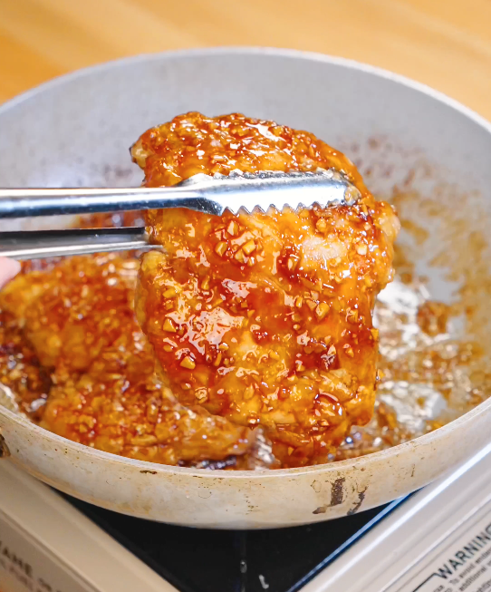 Fried chicken thigh being dipped and coated in thick soy garlic sauce with minced garlic in a pan.