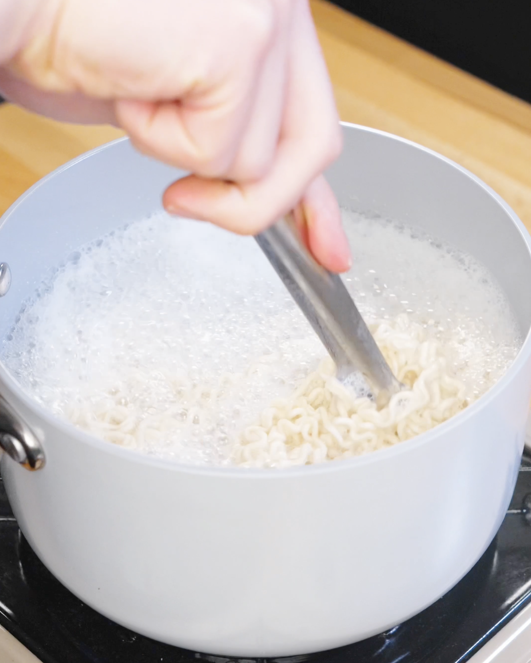 Instant ramen noodles boiling in a pot of water while being stirred with tongs on the stovetop.