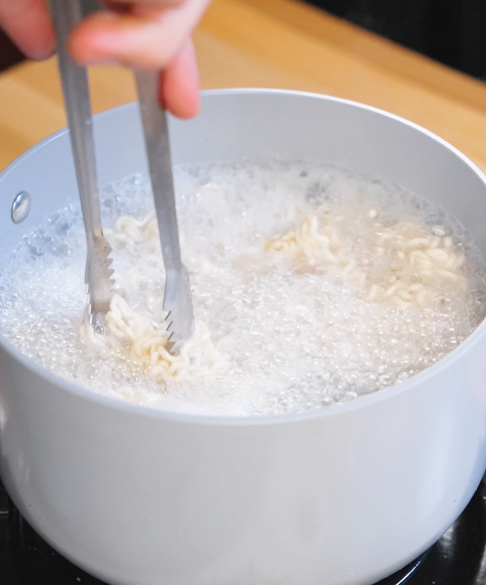 Instant ramen noodles cooking in a pot of boiling water while being separated with tongs for garlic sesame noodles.