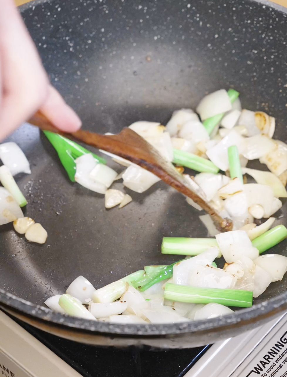 Onion wedges, sliced garlic, and scallion whites saut&eacute;ing in a pan to build the aromatic base for honey garlic chicken stir fry.