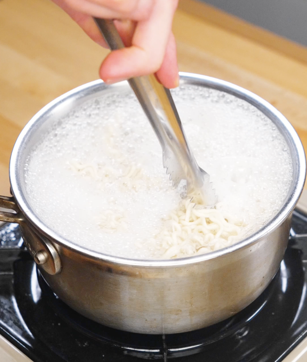 Ramen noodles cooking in a pot of boiling water on the stovetop with tongs stirring.