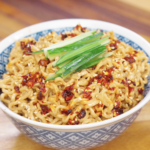 Bowl of creamy sesame noodles topped with chili oil, sesame seeds, and sliced cucumbers.