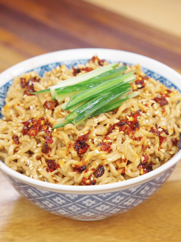 Bowl of creamy sesame noodles topped with chili oil, sesame seeds, and sliced cucumbers.