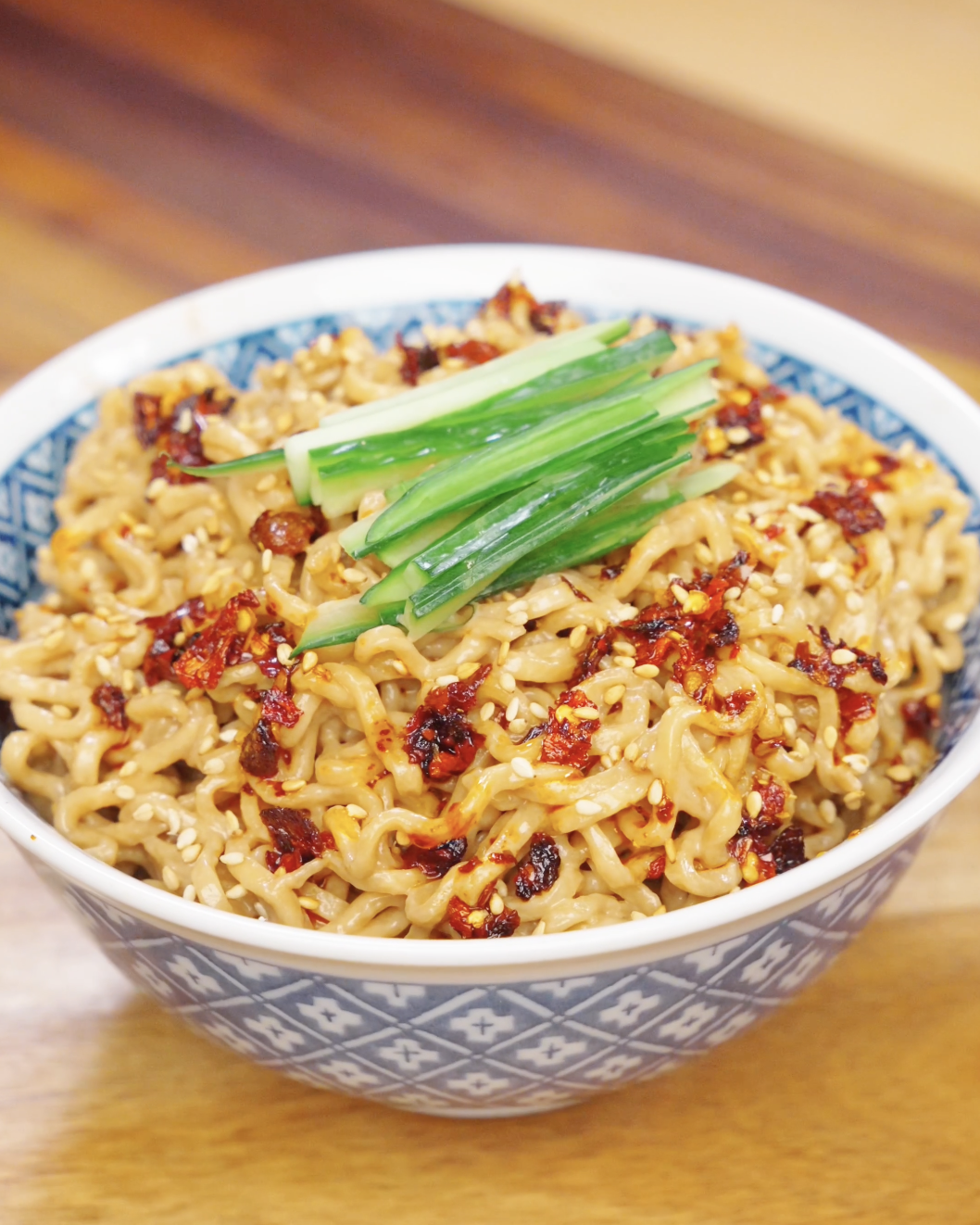 Bowl of creamy sesame noodles topped with chili oil, sesame seeds, and sliced cucumbers.
