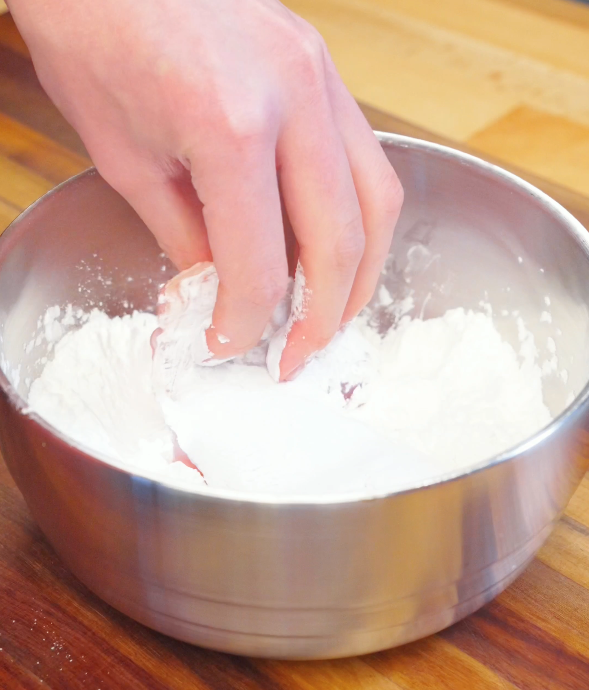 Hand coating raw chicken thighs in cornstarch inside a metal bowl to create a crispy crust for Honey Chili Chicken.