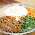 Bowl of beef bibimbap recipe with rice seasoned beef spinach carrots and a fried egg topped with sesame seeds.