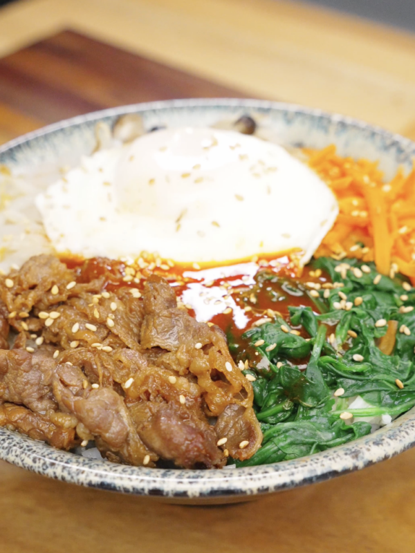Bowl of beef bibimbap recipe with rice seasoned beef spinach carrots and a fried egg topped with sesame seeds.