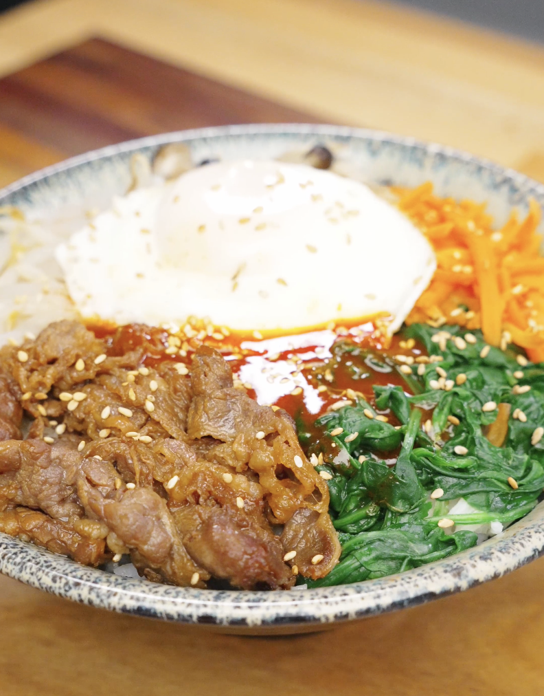 Bowl of beef bibimbap recipe with rice seasoned beef spinach carrots and a fried egg topped with sesame seeds.