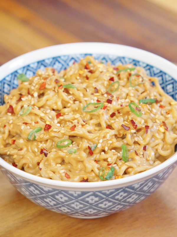 Bowl of creamy peanut noodles garnished with sesame seeds, sliced scallions, and chili flakes.