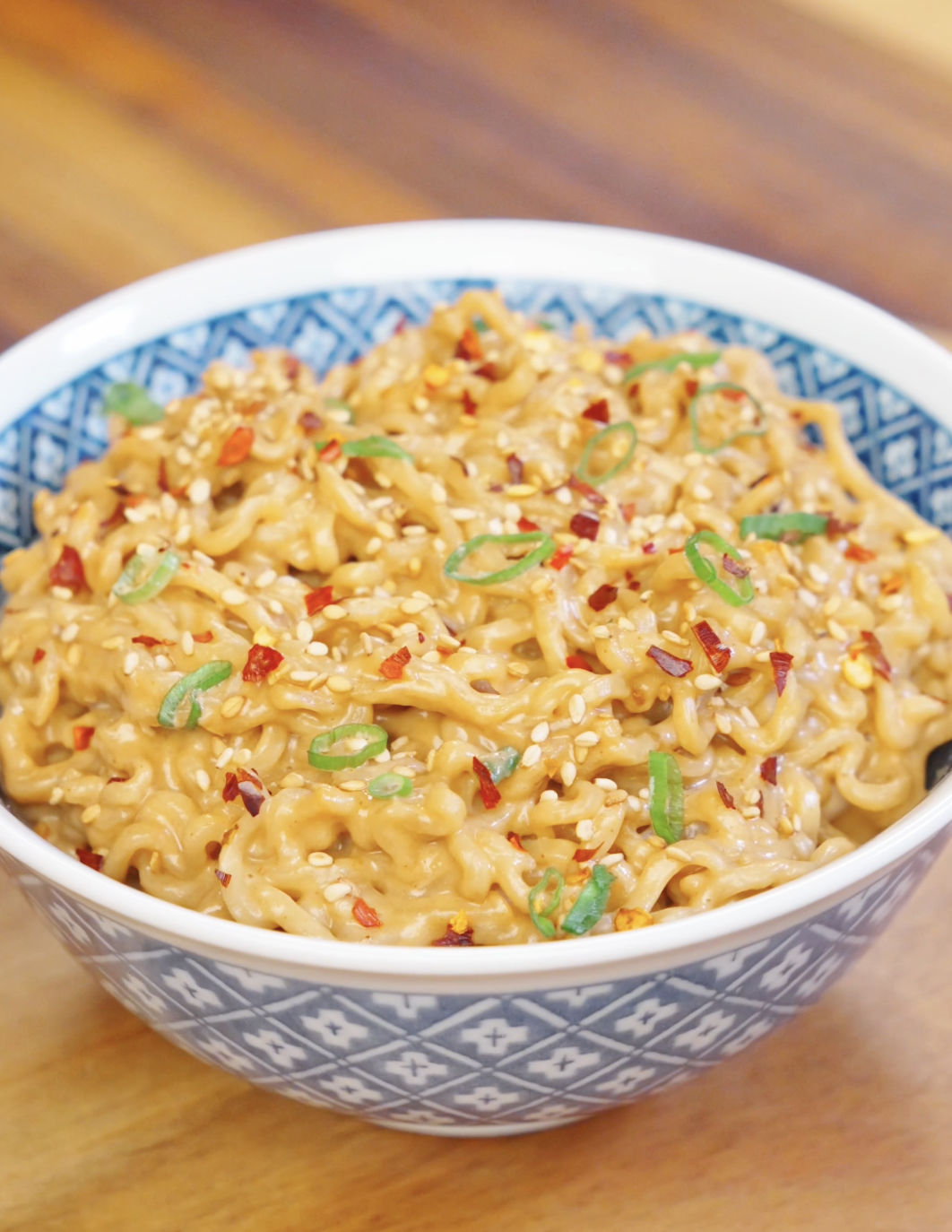 Bowl of creamy peanut noodles garnished with sesame seeds, sliced scallions, and chili flakes.