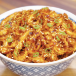 Bowl of glossy garlic chili noodles garnished with sesame seeds, chili flakes, and sliced scallions.