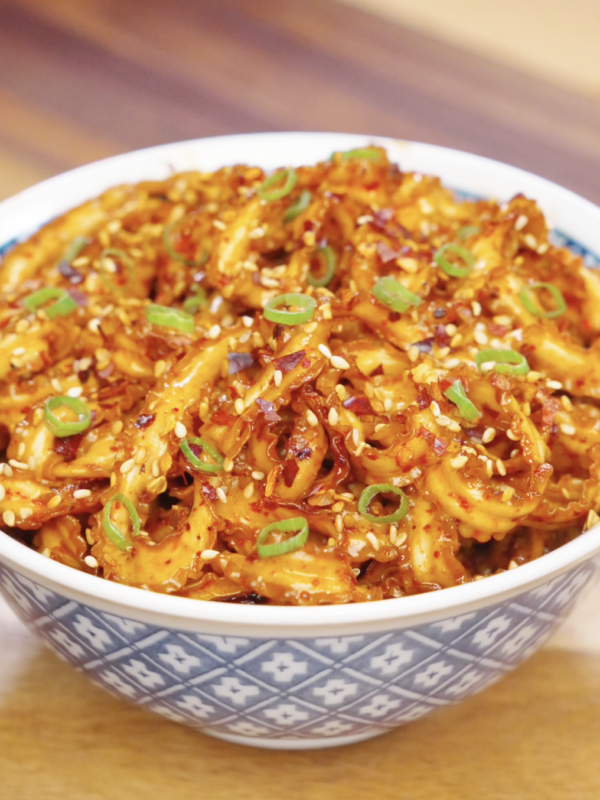 Bowl of glossy garlic chili noodles garnished with sesame seeds, chili flakes, and sliced scallions.