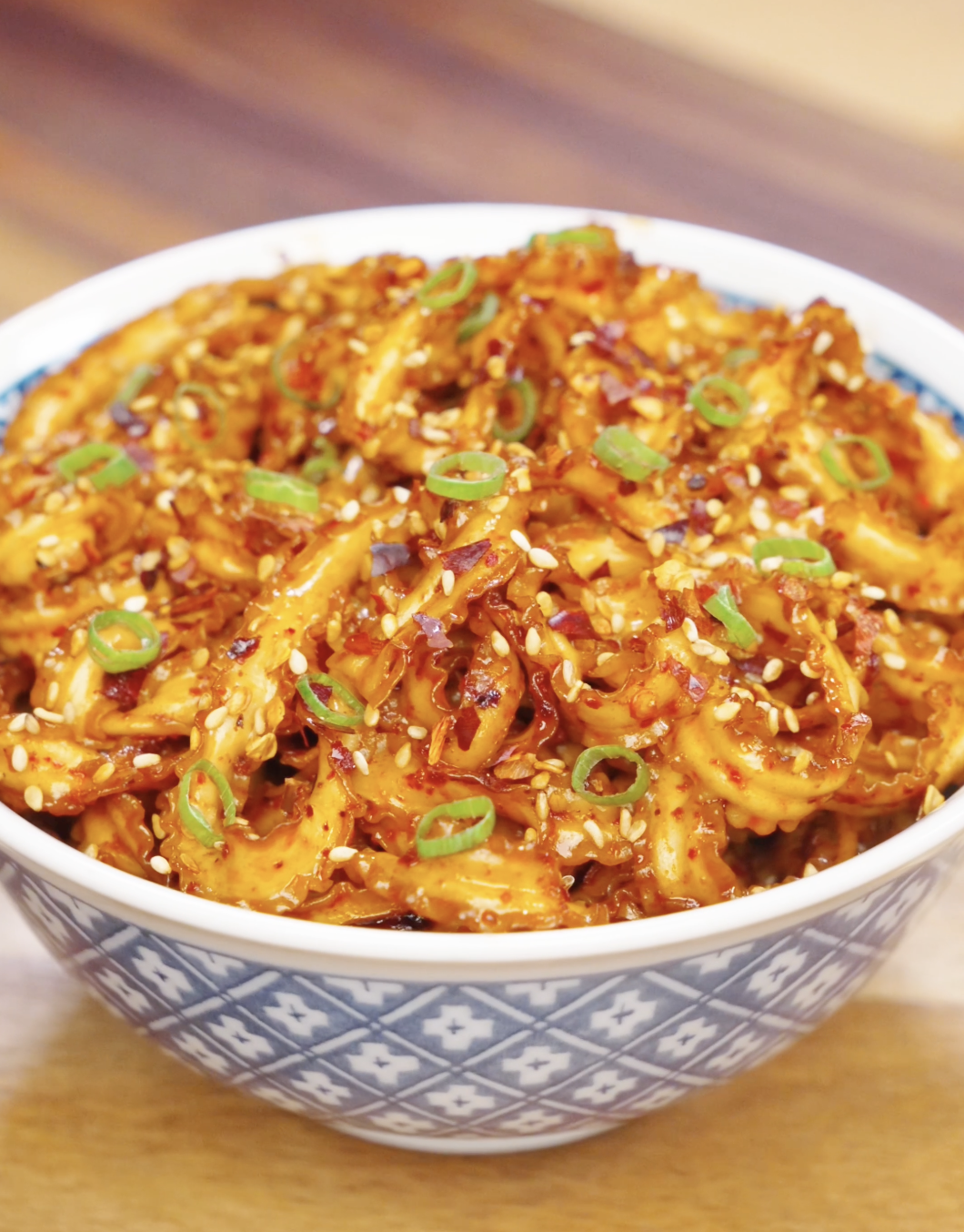 Bowl of glossy garlic chili noodles garnished with sesame seeds, chili flakes, and sliced scallions.