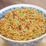 Bowl of garlic peanut butter noodles topped with sesame seeds, chili flakes, and sliced scallions for extra flavor and texture.