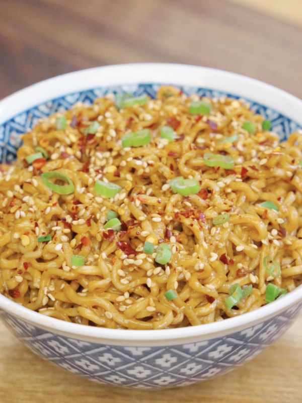 Bowl of garlic peanut butter noodles topped with sesame seeds, chili flakes, and sliced scallions for extra flavor and texture.