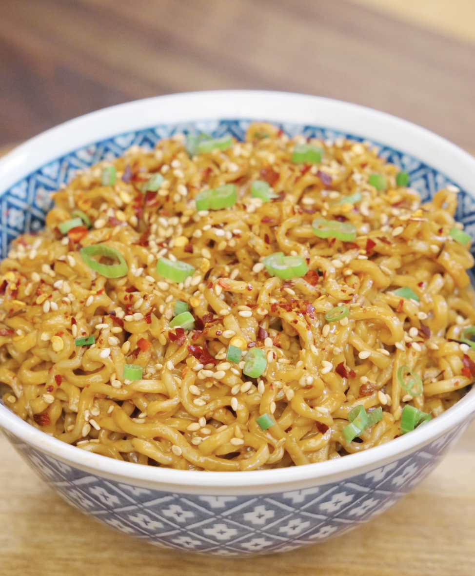 Bowl of garlic peanut butter noodles topped with sesame seeds, chili flakes, and sliced scallions for extra flavor and texture.