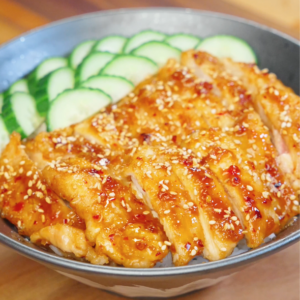 Bowl of sliced Honey Chili Chicken glazed in a sticky sweet and spicy sauce, served over rice with sesame seeds and sliced cucumbers.