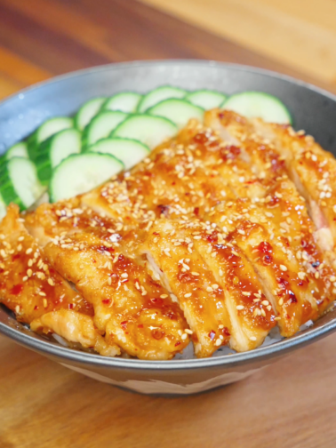 Bowl of sliced Honey Chili Chicken glazed in a sticky sweet and spicy sauce, served over rice with sesame seeds and sliced cucumbers.