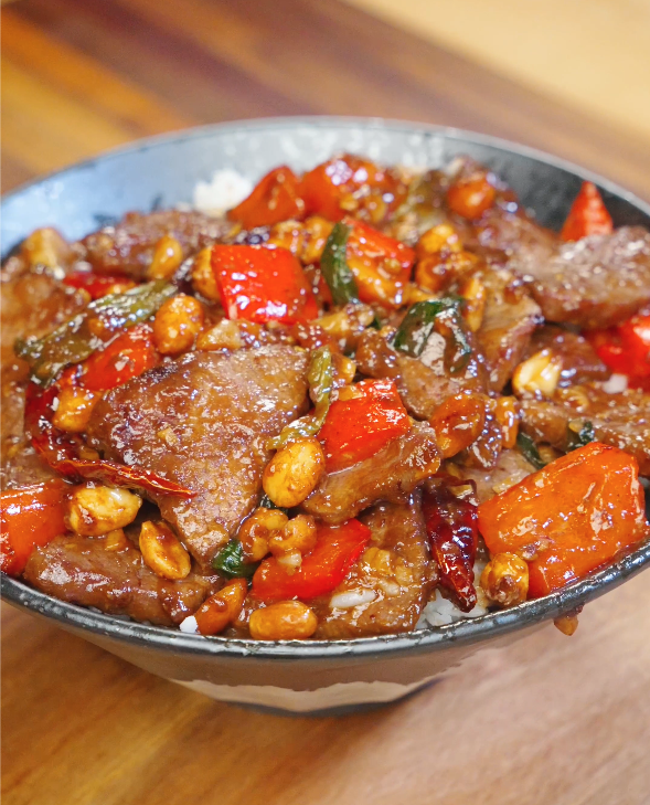 Bowl of Kung Pao Beef served over rice with red bell peppers, dried chilies, scallions, and roasted peanuts coated in kung pao sauce.