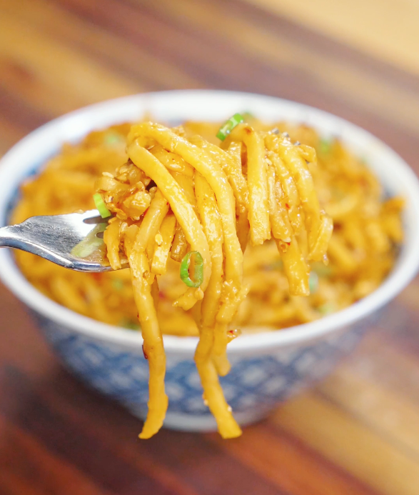 Fork lifting a bundle of chili oil garlic butter noodles coated in buttery chili sauce with sesame seeds and scallions.