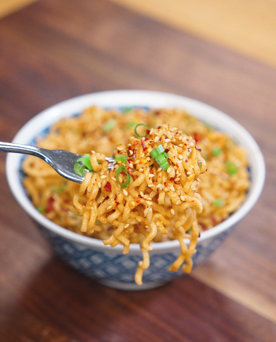 Fork lifting a bite of garlic sesame noodles coated in sesame sauce with chili flakes, sesame seeds, and fresh scallions.